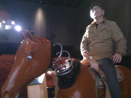 A photo of a man in a theatre, sitting on an artificial brown horse with his head attached by a string to the body.