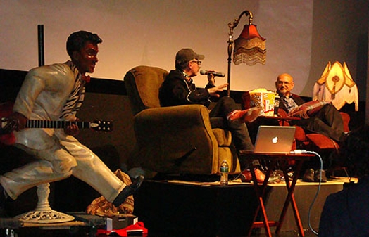 A photo of two people sitting in armchairs and talking on a stage, Near them there is a lamp, some popcorn on a table, a laptop and a white rock star figurine holding a guitar.