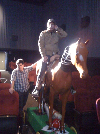 A photo of a man sitting on an artificial brown horse with his head attached by a string to the body. The horse is placed on a green stand with wheels and near it is another person that holds it.