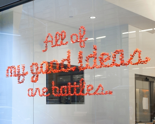 A photo of some orange pieces of plastic on a glass wall forming a text that says: All of my good ideas are battles.