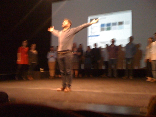 a man dressed in a shirt and jeans is on the stage with hands raised up, and behind them is a line of people