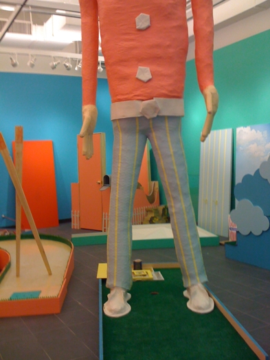 a large statue of a man with torso and legs, with a red shirt and blue trousers with yellow vertical stripes and between the legs is the hole for the mini-golf course
