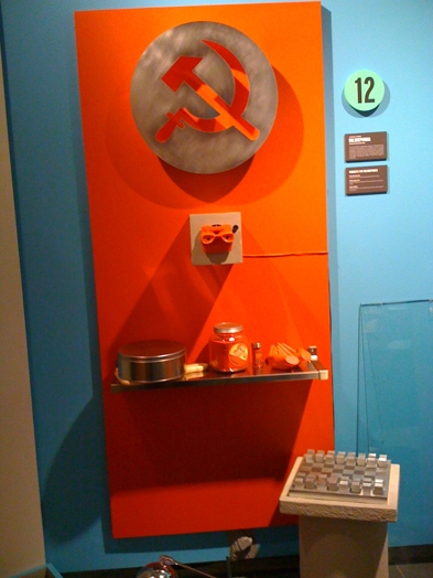 a red shelf with miscellaneous objects on it and a big rounded metal shape with the soviet union symbol