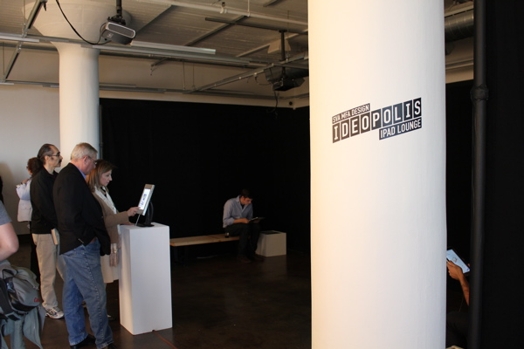 Ideopolist exhibition sticker applied on a column, and to the left side are people gathered near an iPad screen and are looking at it