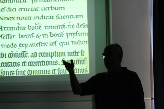 A man giving a lecture while behind him is a screen projector with some old text written in different languages and styles.