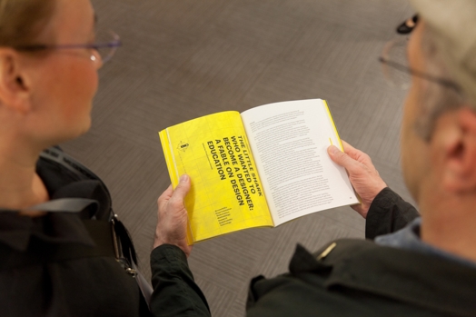 two people looking at an opened book with lots of text paragraphs on a bright yellow page and another white page