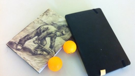 a notebook with an illustration of a working man, a black back cover of a notebook, and two table-tennis yellow balls