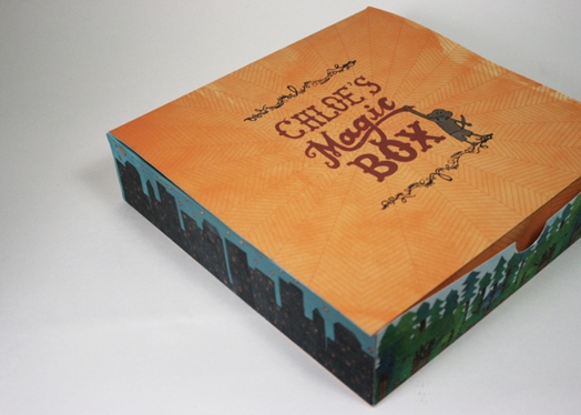 Chloe's magic box with orange lid with a spider web under the logo text, and the sides of the box are with buildings and night sky, a green forest, etc.