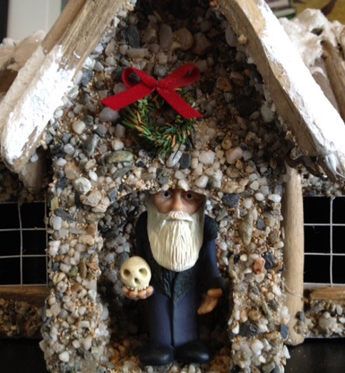 a small figurine of an older man with a white beard in the door of a tiny model house with a red ribbon Christmas decoration above the door