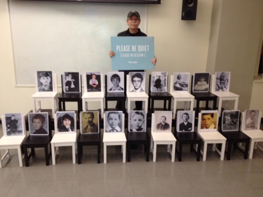 a man standing behind two rows of chairs with portraits on them and the man holds in his hands a sign with "please be quitet"