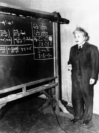 a black and white photo of a man in a black suit near a blackboard with math problems written on it