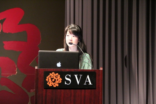 a woman giving a speech from a stand in the "designer as" thesis presentation