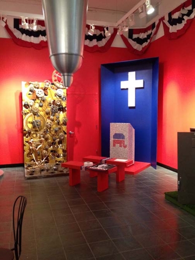 A corner of the room with a white cross inside a blue wooden structure and a tall vertical rectangular with golden cogs and chains