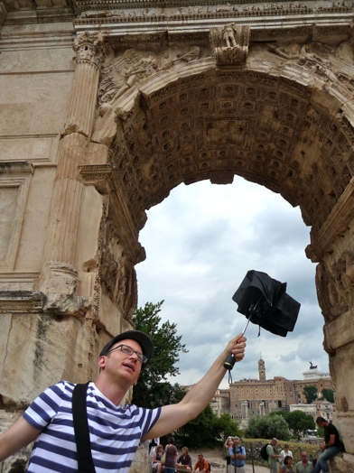 man with glasses holding an umbrella above an arch in Rome