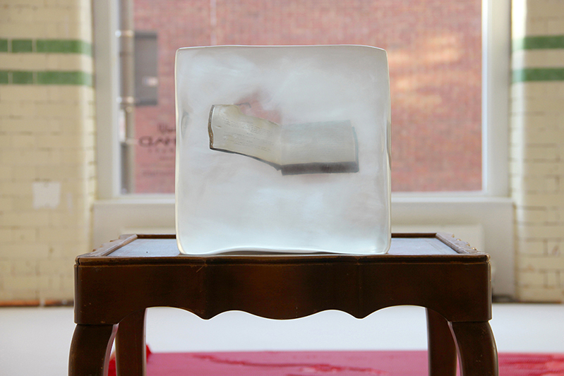 A wooden chair with a transparent acrylic cube with a book in it.