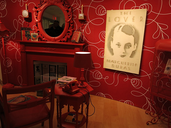 installation view of a red-framed mirror with a showcase cabinet, red furniture, and a white paper poster with a portrait on it, and the title is The Lover
