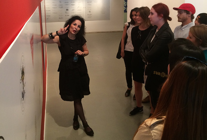A photo of a woman giving a lecture to a group of students while showing some artwork on a wall.