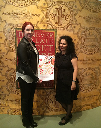 A photo of two women sitting in front of a gold patterned wall and holding a poster with the text: Louise Fili.