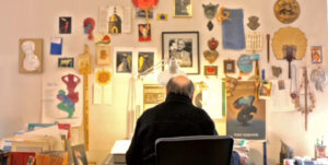 a man working on a desk with the wall in front of him full of artworks
