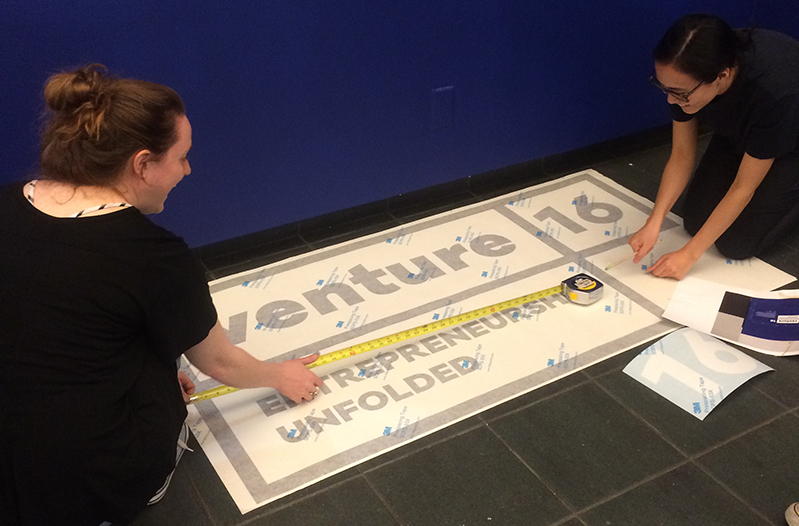 two women are preparing the venture 16 exhibition banner