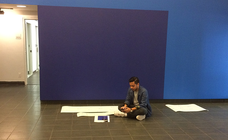 A man preparing some adhesive prints to be put on a blue wall, while sitting in an exhibition room.