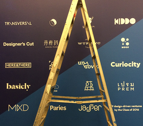 A photo of a metallic golden ladder in front of a black, blue and yellow logo poster on a wall.