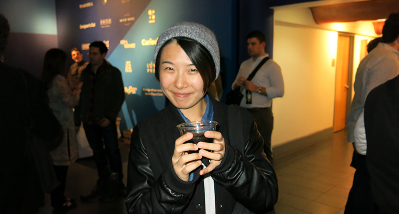 A photo of a smiling woman holding a cup while standing in an exhibition room among other people.