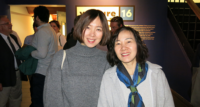 A photo of two women standing in an exhibition room among other people.