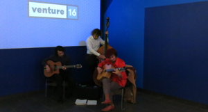 A group of students performing on stage at various musical instruments, while standing in front of a blue projector screen with text logo: venture 16.