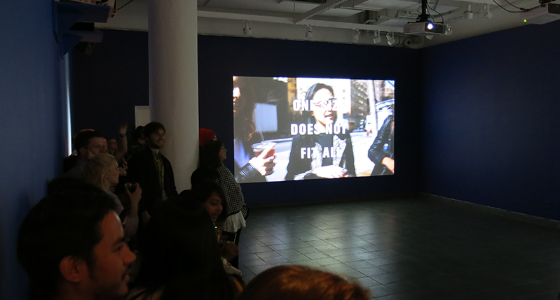 A photo of a group of people standing in an exhibition room while watching a movie presentation on a screen projector.