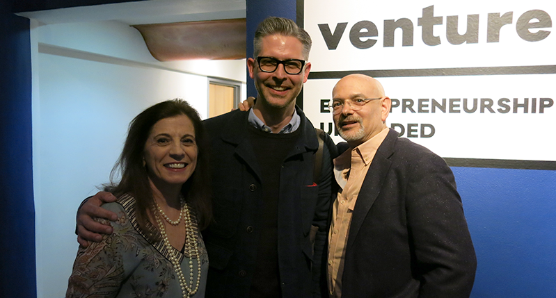 A photo of three people standing in a blue exhibition room with text on walls that says: venture 16 Entrepreneurship Unfolded.