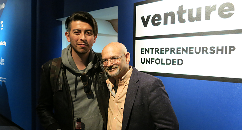 A photo of two people standing in a blue exhibition room with text on walls that says: venture 16 Entrepreneurship Unfolded.