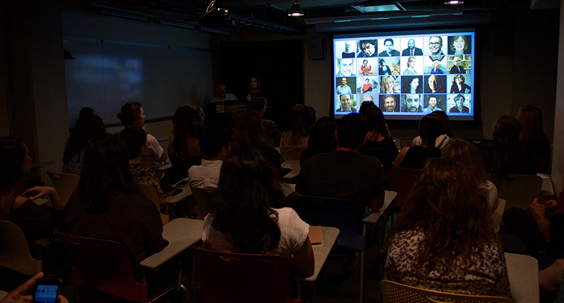 A group of people sitting in a classroom and watching a conference of multiple people on a screen projector.