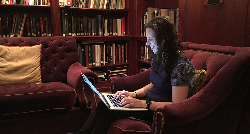 A photo of a woman sitting on an armchair near a couch and a bookshelf, while working on a laptop.
