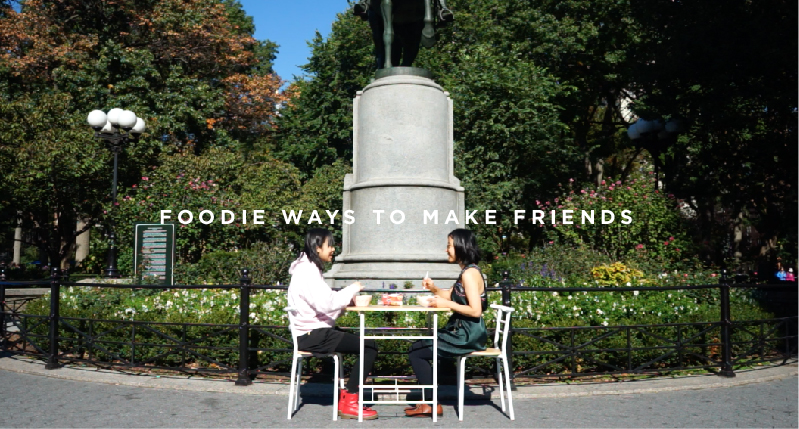 A photo of two girls sitting at a table in a park, while eating something. On the photo there is the text: Foodie Ways To Make Friends.