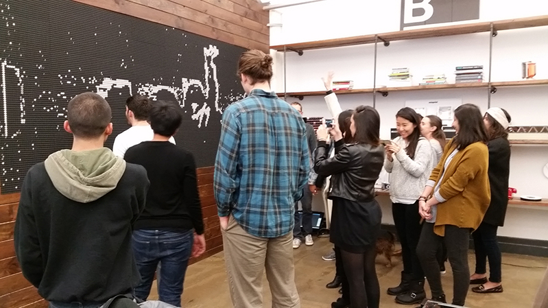 A photo of a group of students standing in an exhibition room, while looking at a blackboard with some white drawing.