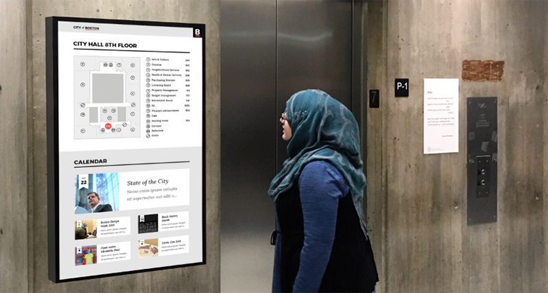 A photo of a woman wearing a blue blouse and a scarf, standing near an elevator door while looking at a building plan schematic.