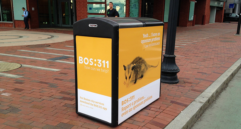 A street mailbox in the street with a yellow and white poster on it. The poster shows an image of an animal and some text.