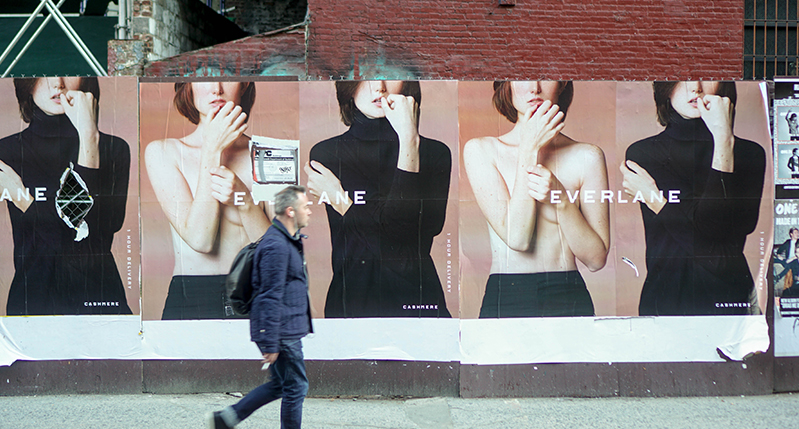 a person passing by on the sidewalk and on the building behind him are posters of everlane