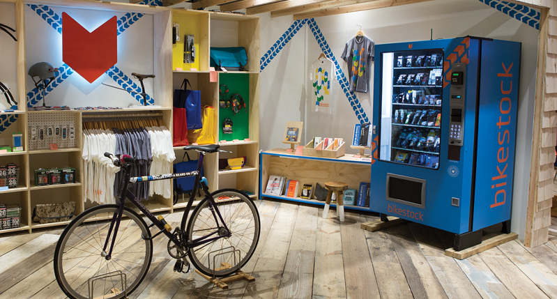 A photo of a room with a bicycle in the middle and around, near each wall there are some shelves with different items like t-shirts, bags, books and other colorful items.