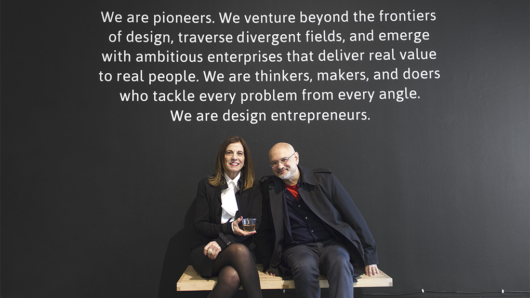A photo of two teachers sitting on a bench. Also behind them on a wall there is a text: We are pioneers. We venture beyond the frontiers of design, travers divergent fields, and emerge with ambitious enterprises that deliver real value to real people. We are thinkers, makers, and doers who tackle every problem from every angle. We are design entrepreneurs.