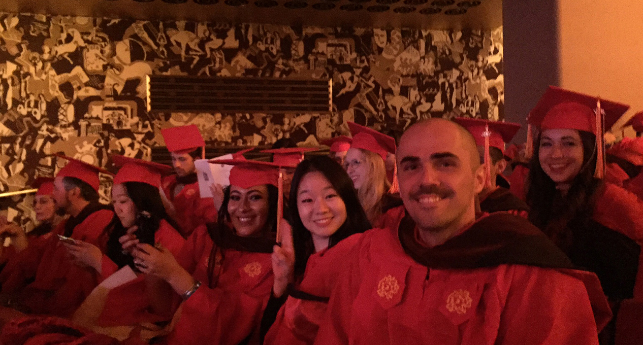 A photo of a group of grad students dressed in SVA colored graduation clothes while sitting in a theatre room.