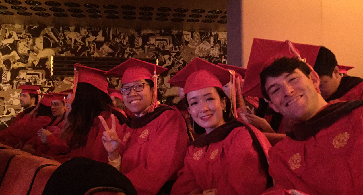 A photo of a group of grad students dressed in SVA colored graduation clothes while sitting in a theatre room.