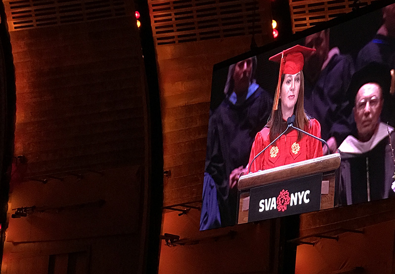 A photo of a tv screen in a theatre room. On it there is a student from SVA NYC speaking to the audience on graduation day.