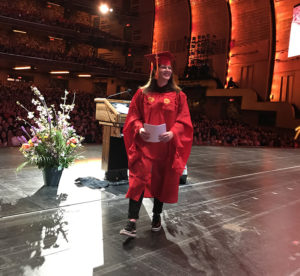 A photo of a woman dressed in SVA graduation clothes, that is walking on the stage in front of the audience with some piece of paper in her hand.