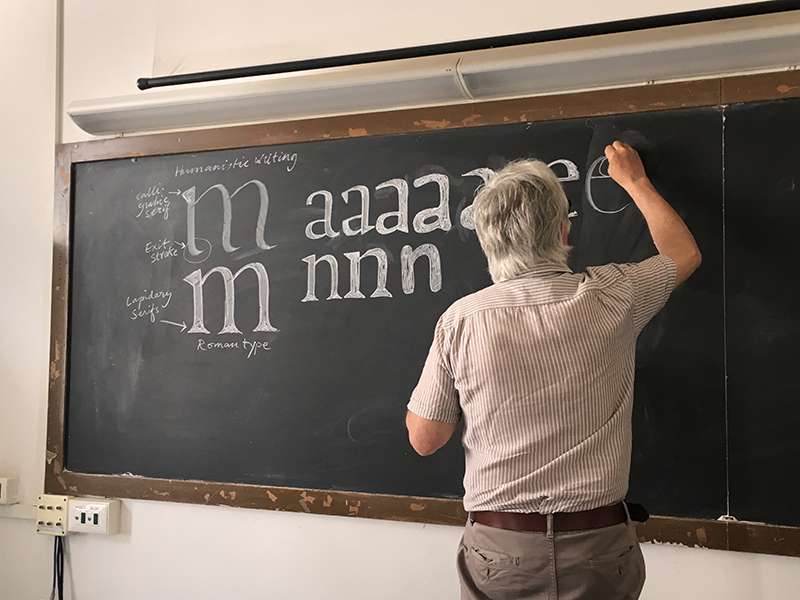 A man at a blackboard, teaching the typography and art of drawing letters.