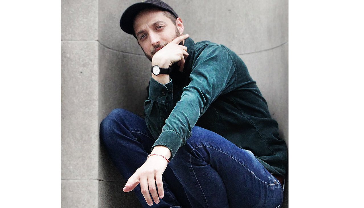 A man with a watch and cap, near a wall thinking and pointing a finger.