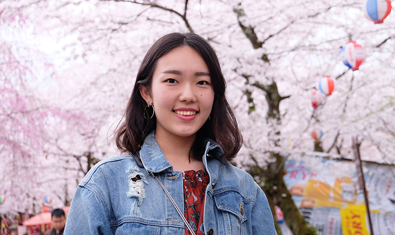 A smiling girl and in the background some pink blossomed trees.