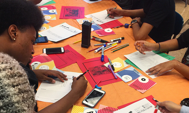 A photo of a group of students siting at a table and doing some sketches on pieces of colored paper. On the table there is also flyers, crayons and mobile phones.