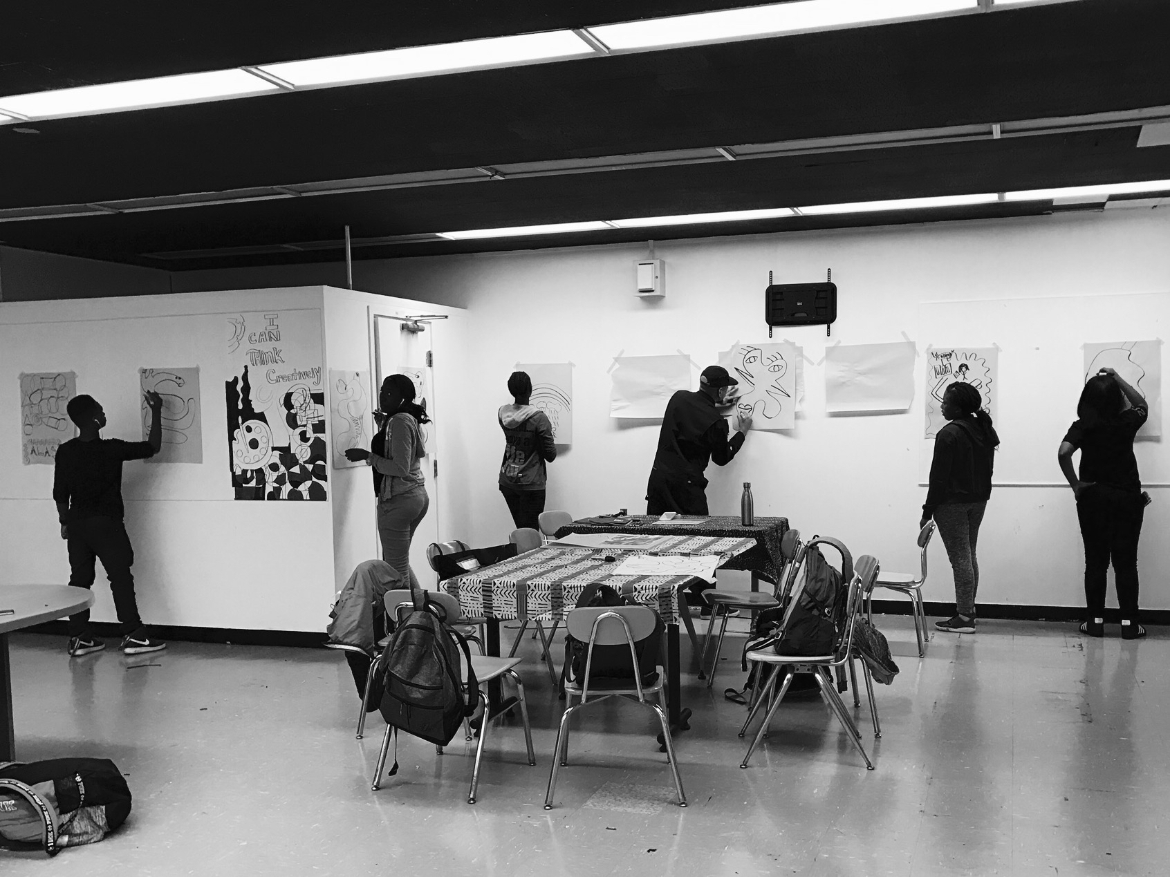 A black and white photo of an art exhibition where people are drawing on sheets of paper glued to the wall. Also in the middle of the room there is an empty table with chairs around it. On each chair there is a backpack.